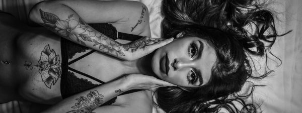 Tattooed escort laying face up with hands against her cheeks