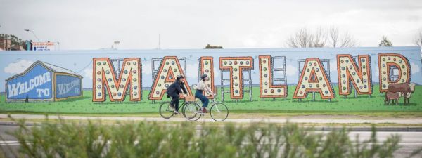 A couple riding bicycles on a path in front of the 'welcome to Maitland' NSW sign